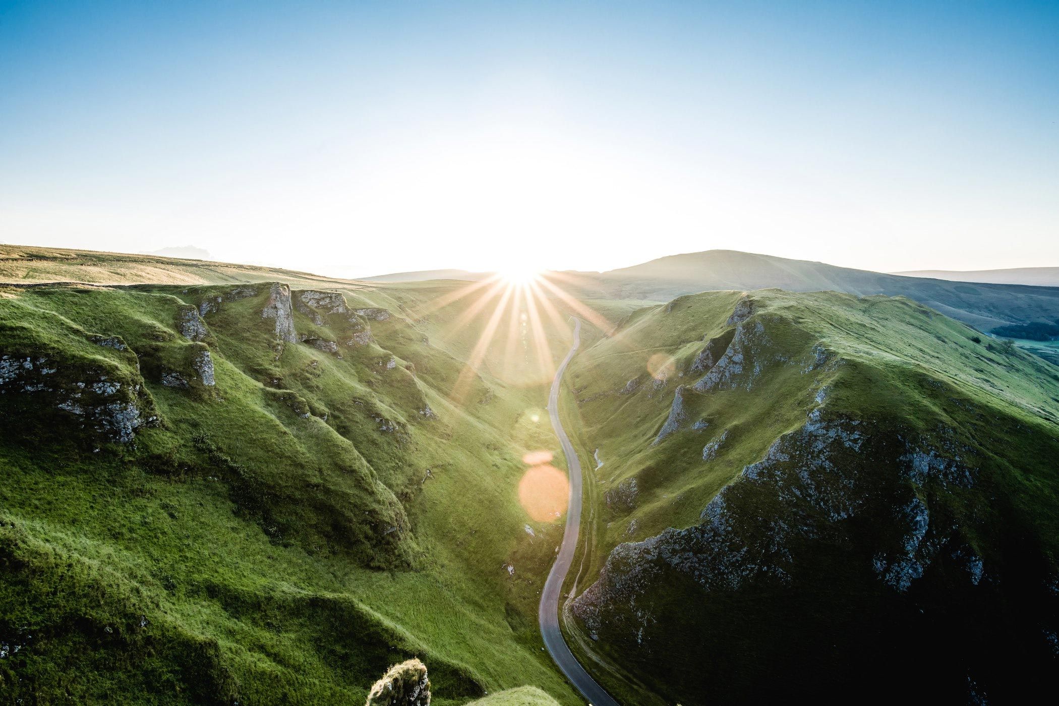 aerial landscape shot with road in the middle of large green hills leading towards sun rising over horizon